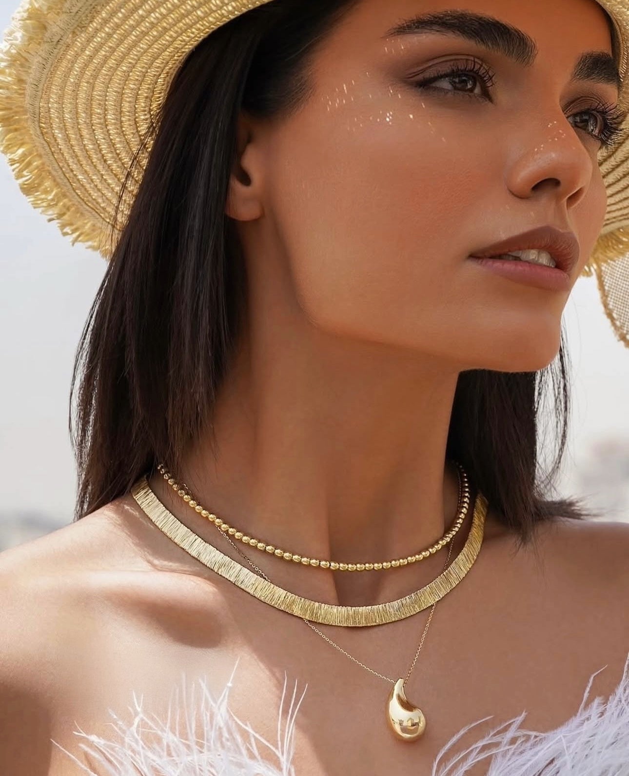 Nahaufnahme einer Frau mit Strohhut, die eine elegante 18k Gelbgold-Perlenkette dicht am Hals trÀgt, kombiniert mit einer breiten, strukturierten Goldcollier-Kette und einer feinen Kette mit tropfenförmigem AnhÀnger.