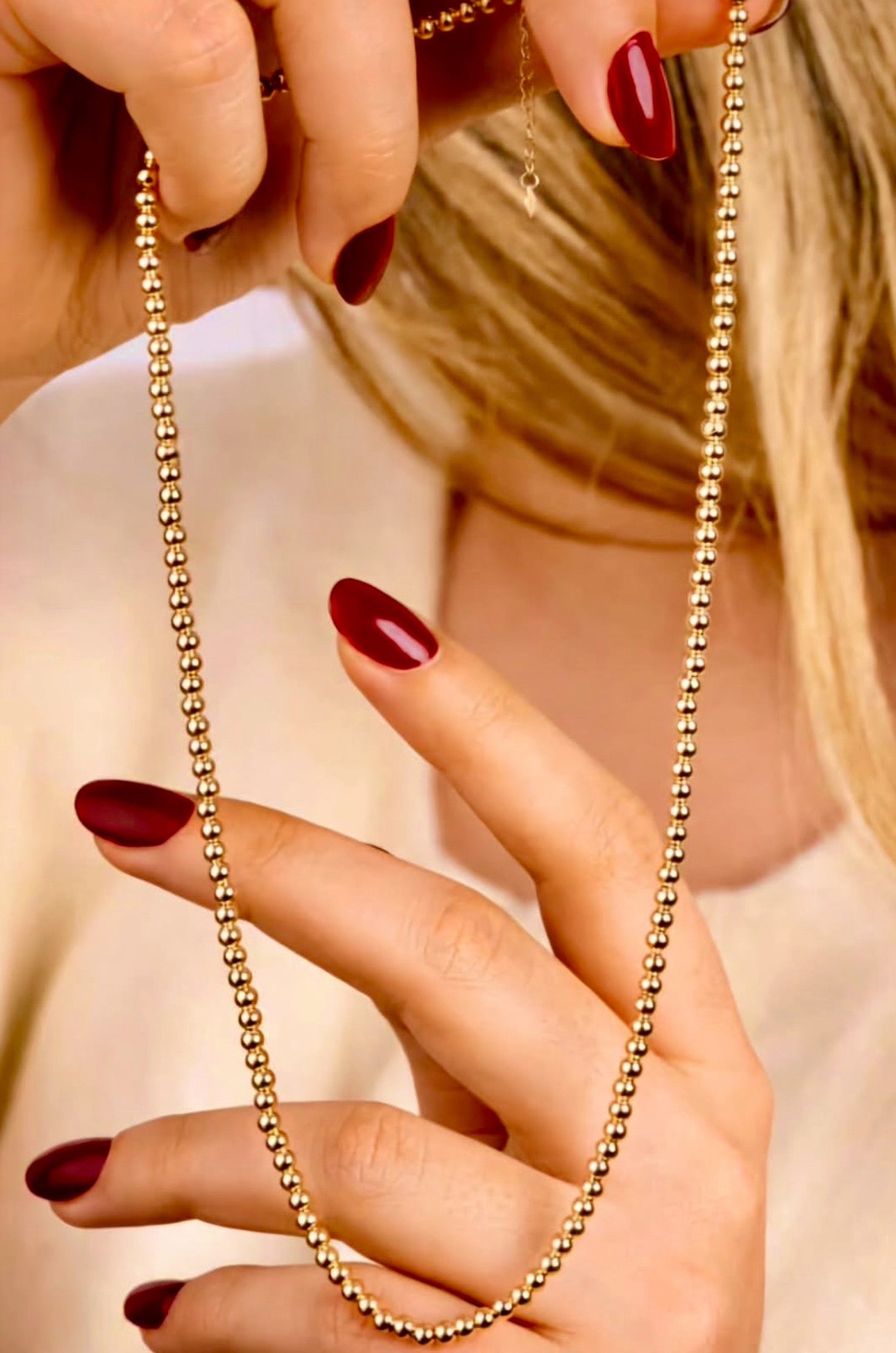 Nahaufnahme von HĂ€nden mit dunkelrotem Nagellack, die eine 18k Gelbgold-Perlenkette mit gleichmĂ€Ăig angeordneten, polierten Goldkugeln halten, vor einem weichen, beigefarbenen Hintergrund.