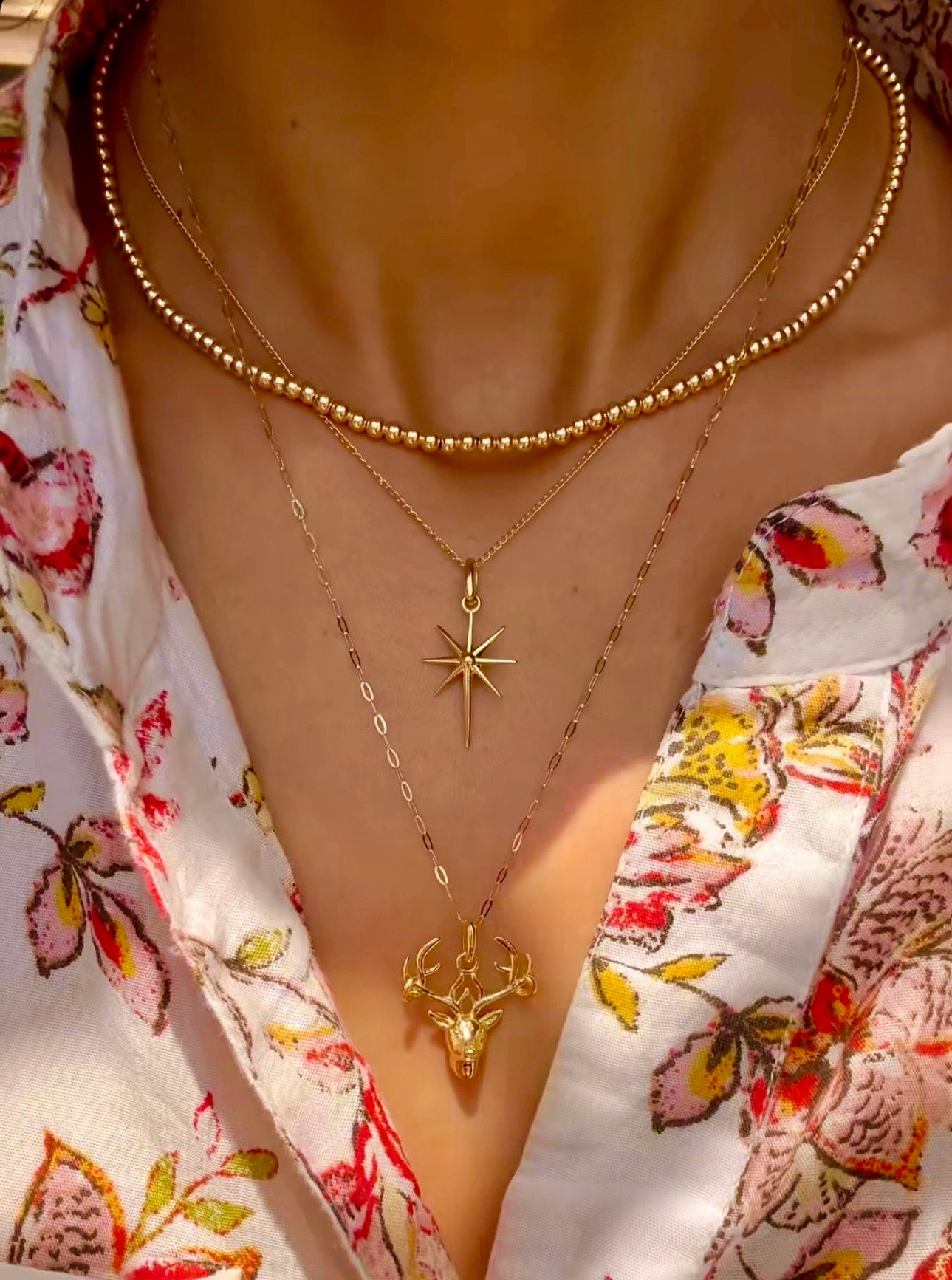 Nahaufnahme eines Halses mit drei ĂŒbereinander getragenen Halsketten aus 18k Gelbgold. Die oberste Kette ist eine feine Perlenkette mit gleichmĂ€Ăig runden, polierten Goldkugeln. Darunter hĂ€ngt eine filigrane Kette mit einem achtzackigen SternanhĂ€nger, und die lĂ€ngste Kette trĂ€gt einen detailreichen Hirschkopf-AnhĂ€nger. Getragen zu einer weiĂen Bluse mit farbigem Blumenmuster.