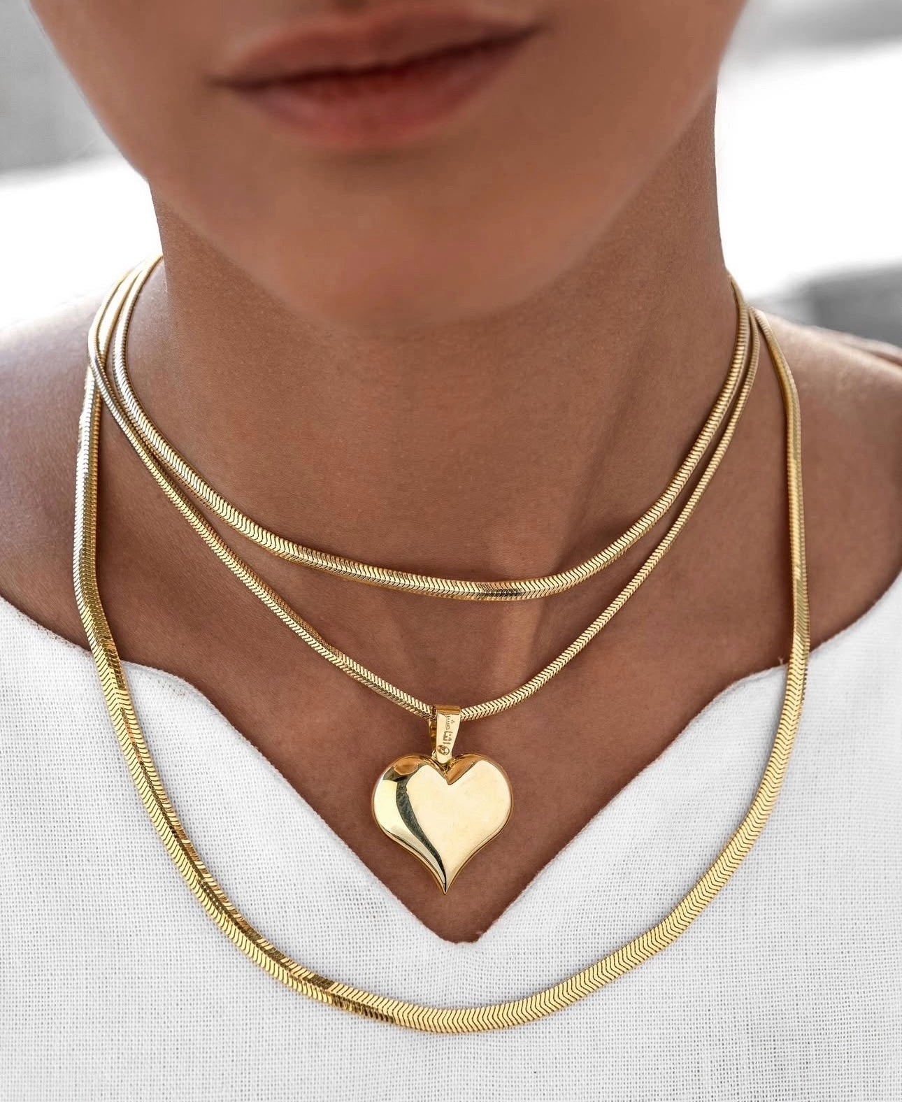 Nahaufnahme eines Halses mit drei übereinander getragenen Schlangenketten aus 18k Gelbgold. Die mittlere Kette ist mit einem polierten Herzanhänger versehen, dessen asymmetrische Form einen modernen, fließenden Look erzeugt. Getragen zu einem weißen, leichten Oberteil mit V-Ausschnitt.
