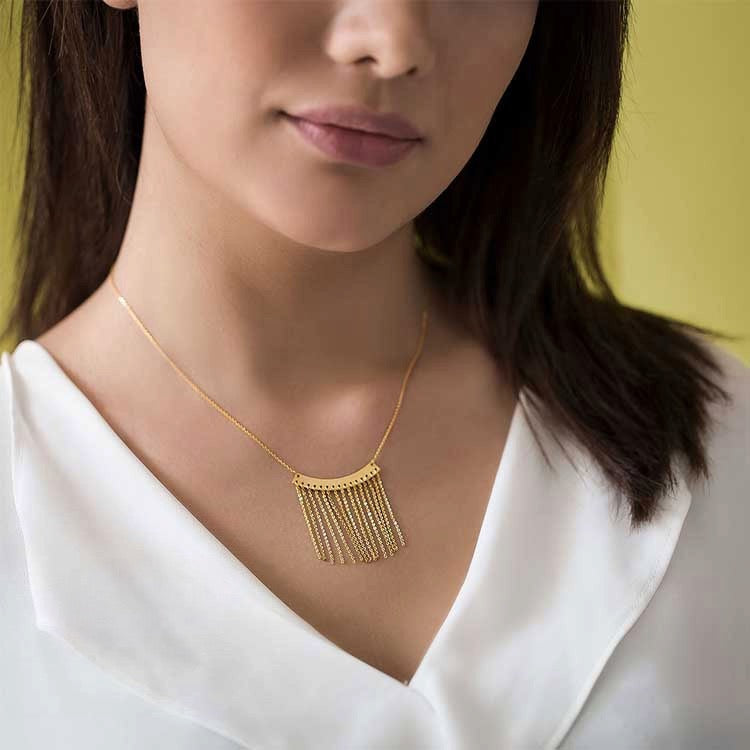 Nahaufnahme einer Frau mit einer Halskette aus 18k Gelbgold. Der Anhänger „Baran-e Zarrin“ (Goldener Regen) besteht aus einer gebogenen Goldplatte, von der mehrere feine Goldketten wie Regentropfen herabhängen. Getragen zu einer eleganten weißen Bluse vor gelbem Hintergrund.
