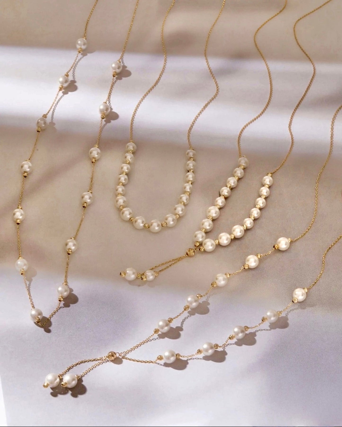 Elegante Halsketten aus 18k Gelbgold mit weißen Perlen, in verschiedenen Längen und Anordnungen, die Perlen wie tanzend auf den feinen Goldketten verteilt, sanft im Licht schimmernd.
