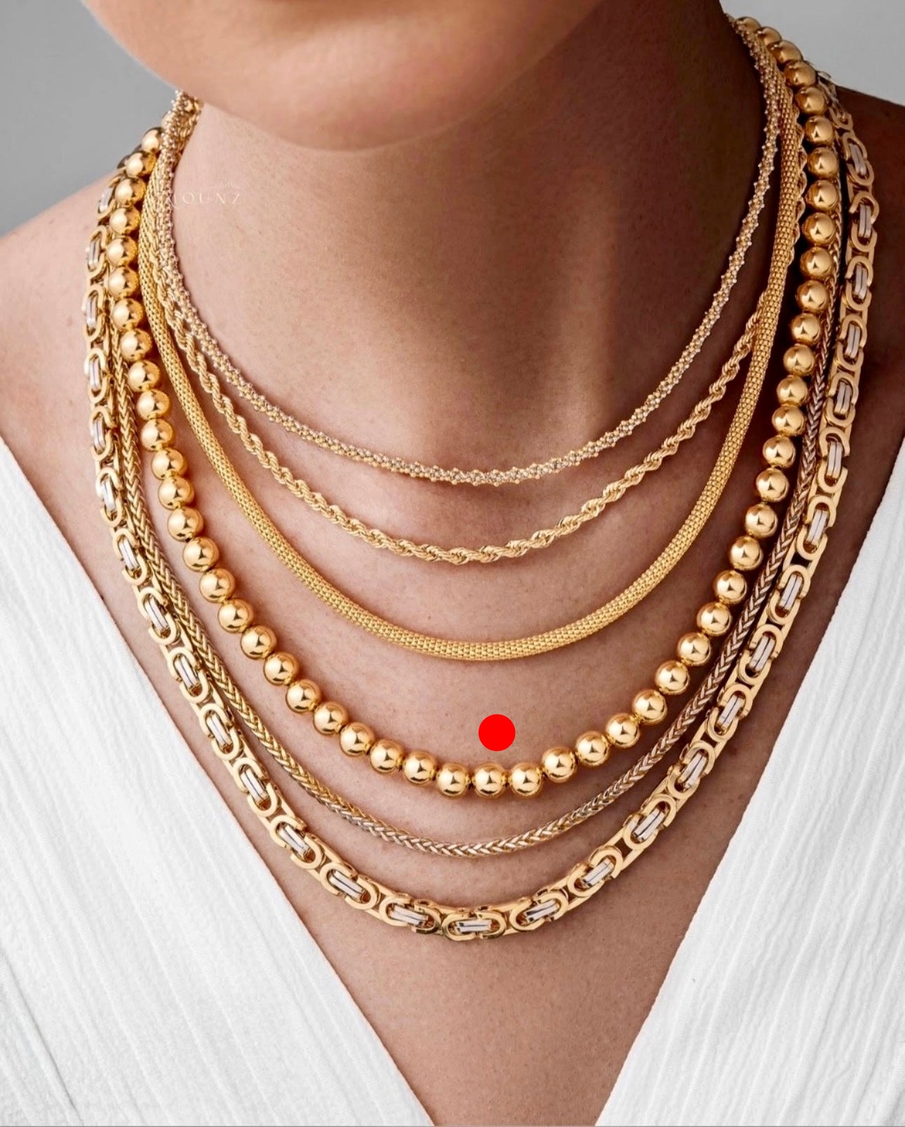 Goldperlenkette aus 18k Gelbgold im Layering-Look getragen, kombiniert mit weiteren Ketten auf heller Haut und weißem Oberteil, markiert für Fokus