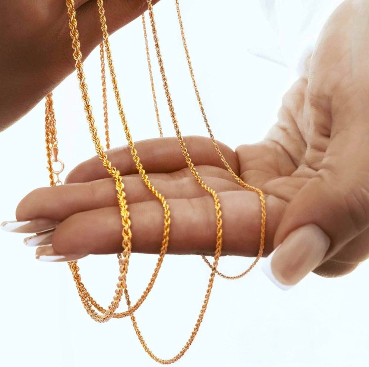 Nahaufnahme einer filigranen Kordelkette aus 18k Gelbgold, die locker über eine geöffnete Hand mit gepflegten, langen Fingernägeln gelegt ist. Die feine, verdrehte Gliederstruktur reflektiert das Licht und betont den warmen Goldglanz. Hintergrund unscharf in hellem Weiß gehalten.
