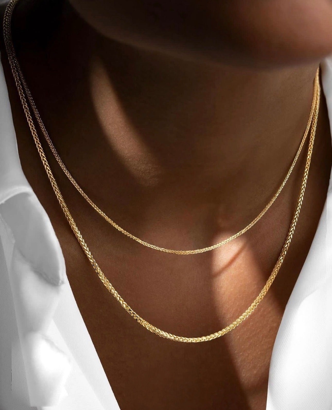 Nahaufnahme eines Halses mit zwei filigranen, glänzenden Miroketten aus 18k Gelbgold und Weissgold in feiner Flechtstruktur, getragen über einem weißen, leicht geöffneten Hemd.