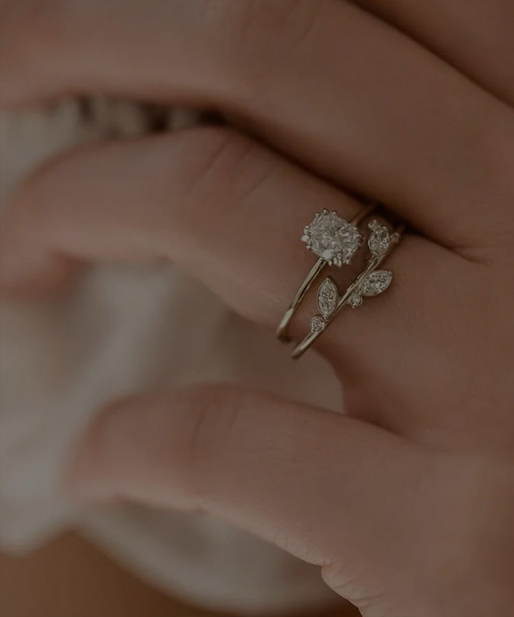 Hand mit zwei filigranen Ringen am Ringfinger: einer mit einem Diamantsolitär, der andere einer Pflanzenranke nachempfunden, die Blättchen mit kleinen Diamanten besetzt.