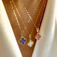 Anhänger in vierblättriger Form aus rosa Stein, eingefasst in feine goldene Perlenfassung, an einer zarten Goldkette, elegant auf heller Stoffbluse getragen.