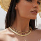 Nahaufnahme einer Frau mit Strohhut, die eine elegante 18k Gelbgold-Perlenkette dicht am Hals trägt, kombiniert mit einer breiten, strukturierten Goldcollier-Kette und einer feinen Kette mit tropfenförmigem Anhänger.