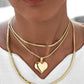 Nahaufnahme eines Halses mit drei übereinander getragenen Schlangenketten aus 18k Gelbgold. Die mittlere Kette ist mit einem polierten Herzanhänger versehen, dessen asymmetrische Form einen modernen, fließenden Look erzeugt. Getragen zu einem weißen, leichten Oberteil mit V-Ausschnitt.