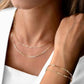 Nahaufnahme einer Halskette aus 18k Gelbgold („Reshteh-ye Sadeh“ – Schlichtes Band) mit länglichen, rechteckigen Gliedern, die locker um den Hals liegen. Passend dazu ein Armband im identischen Design, das doppelt um das Handgelenk gewickelt ist. Getragen zu einem weißen, schulterfreien Oberteil und weißem Blazer.