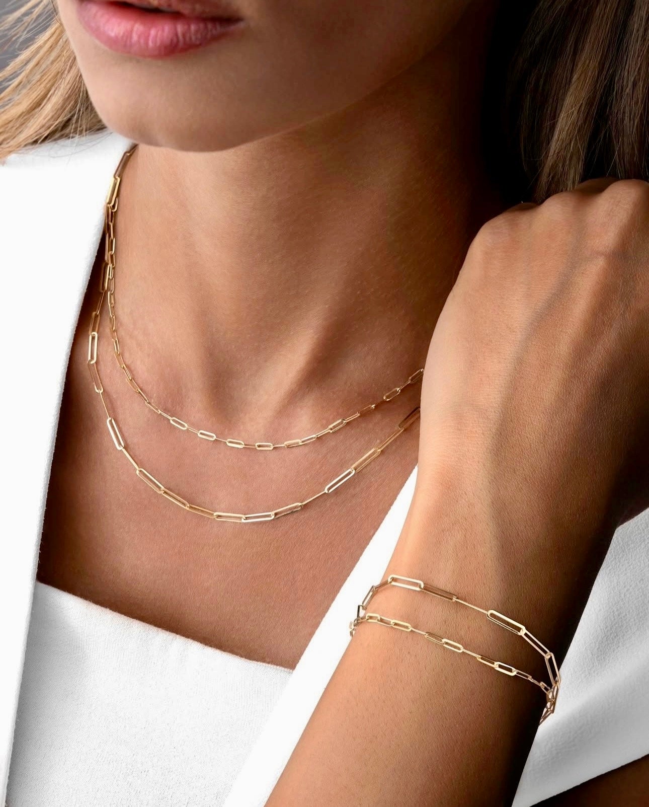 Nahaufnahme einer Halskette aus 18k Gelbgold („Reshteh-ye Sadeh“ – Schlichtes Band) mit länglichen, rechteckigen Gliedern, die locker um den Hals liegen. Passend dazu ein Armband im identischen Design, das doppelt um das Handgelenk gewickelt ist. Getragen zu einem weißen, schulterfreien Oberteil und weißem Blazer.