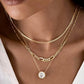 Nahaufnahme eines Halses mit drei übereinander getragenen Halsketten aus 18k Gelbgold. Die mittlere Kette ist eine konische Figaro-Kette mit wechselnden langen und kurzen Gliedern. Darüber liegt eine feine, engmaschige Kette, darunter eine filigrane Kette mit einem großen, ovalen Anhänger, besetzt mit einem funkelnden, klaren Stein. Getragen zu einem tief ausgeschnittenen, weißen Oberteil.