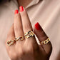Nahaufnahme einer Hand mit rot lackierten Nägeln, die einen eleganten 18k Gelbgold-Ring in Knotenform trägt, Teil eines modernen Ringensembles, vor einem weichen, beigefarbenen Hintergrund.