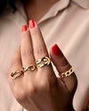 Nahaufnahme einer Hand mit rot lackierten Nägeln, die einen eleganten 18k Gelbgold-Ring in Knotenform trägt, Teil eines modernen Ringensembles, vor einem weichen, beigefarbenen Hintergrund.